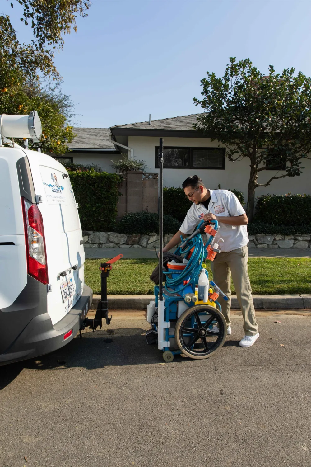 Uniformed pool technician unloading an equipment cart from a branded service van parked on a residential street.