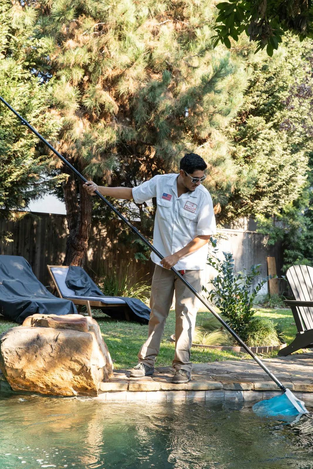 Person in light-colored shirt using long-handled skimmer net to clean pool water, with large rocks and green plants visible.