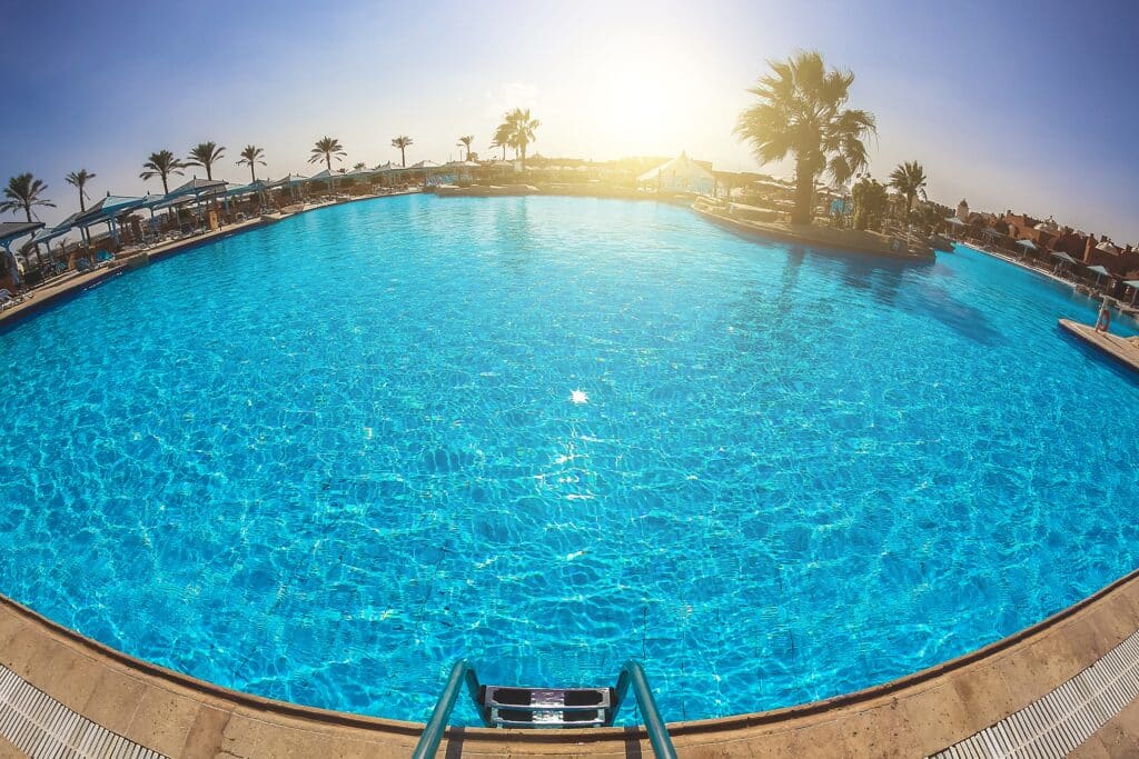 Large, clear blue swimming pool with sun reflecting off the water, surrounded by palm trees and lounge chairs under a bright sky in Long Beach CA. Poolside steps are visible in the foreground, perfect for enjoying quality pool service.