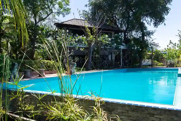 A clear blue swimming pool designed by a skilled swimming pool contractor is surrounded by lush greenery, with a two-story house featuring a balcony and tiled roof in the background under a sunny sky.