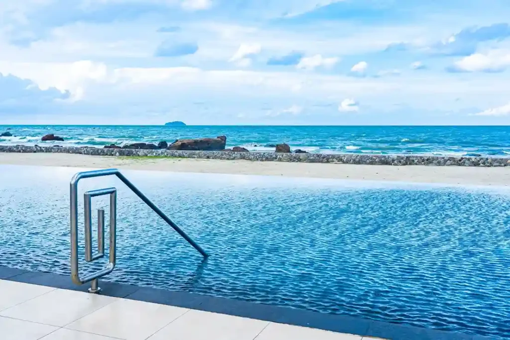 A serene swimming pool overlooks a sandy beach and the blue ocean under a partly cloudy sky, with metal pool steps in the foreground and rocks along the shoreline—perfect inspiration for your next pool service in Long Beach CA.