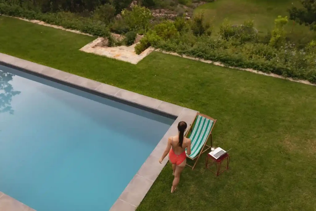 A woman in a red swimsuit walks toward a striped lounge chair and small table with a laptop, beside a sparkling commercial lap pool installation in a grassy backyard.