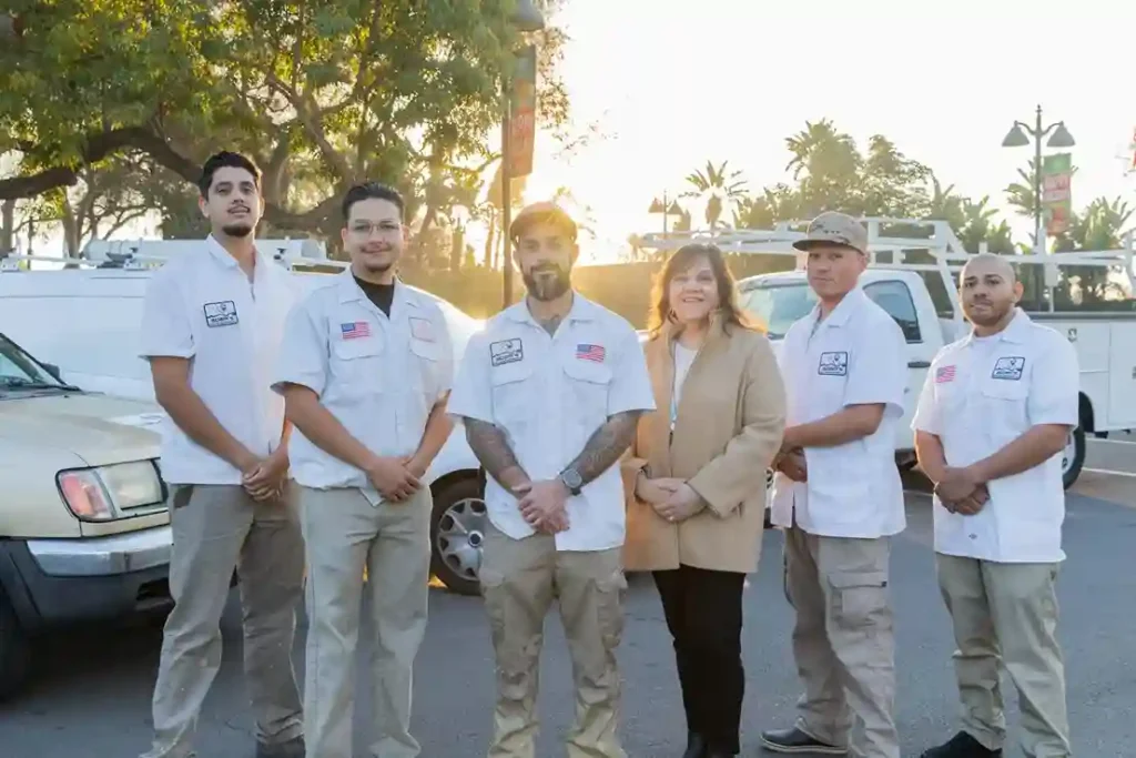 Six people stand outdoors in front of work trucks, representing a Commercial Spa Installation team; five men wear matching white uniforms and one woman in a tan blazer joins them. Trees and sunlight fill the background.