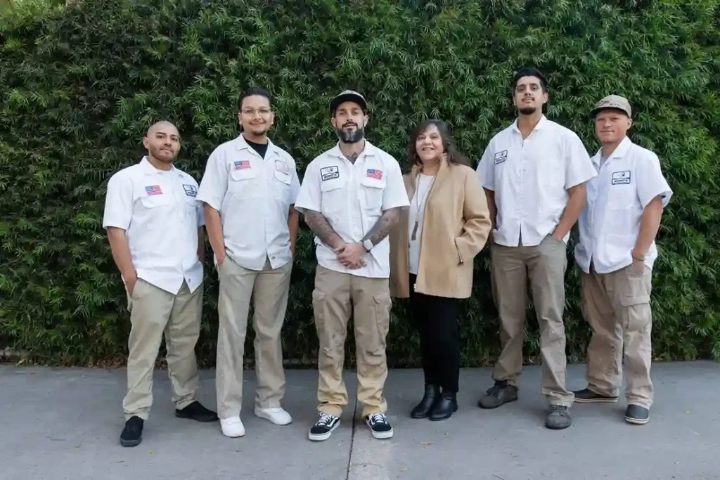 Six people stand in a row outside in front of green bushes. Five men in matching white work shirts and khaki pants and a woman in a tan coat and black pants smile at the camera, representing a Commercial Spa Installation team.