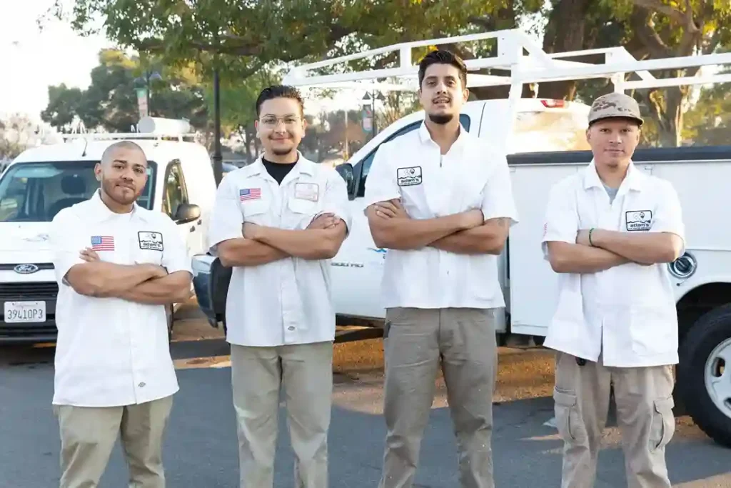 Four men stand outdoors with arms crossed, wearing uniforms of white shirts and khaki pants. Two white work trucks, used for commercial spa installation, are parked behind them, with trees visible in the background.