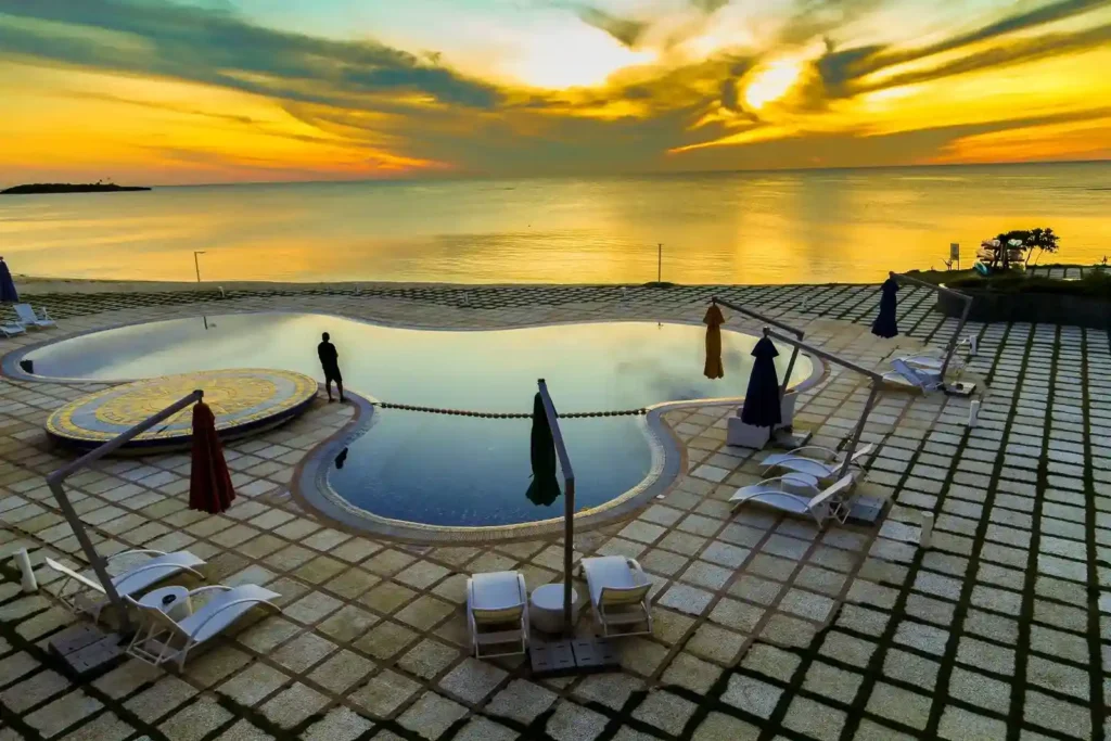 A person stands near a pool crafted by a Residential Swimming Pool Contractor, surrounded by lounge chairs and closed umbrellas, overlooking a calm sea at sunset with dramatic clouds above.