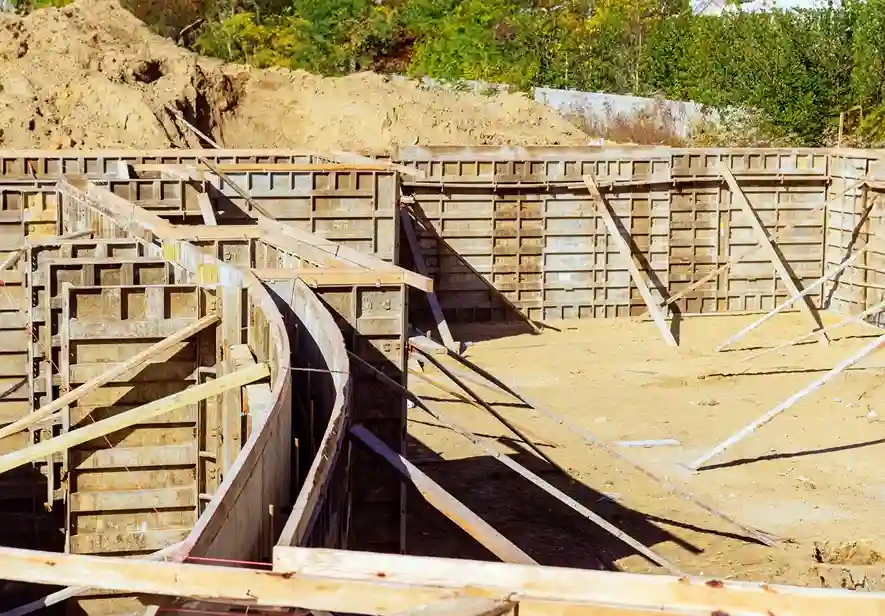 Wooden formwork for a concrete foundation at a construction site, with curved and straight sections on sandy ground—ideal for a residential swimming pool contractor—surrounded by vegetation and a dirt mound in the background.