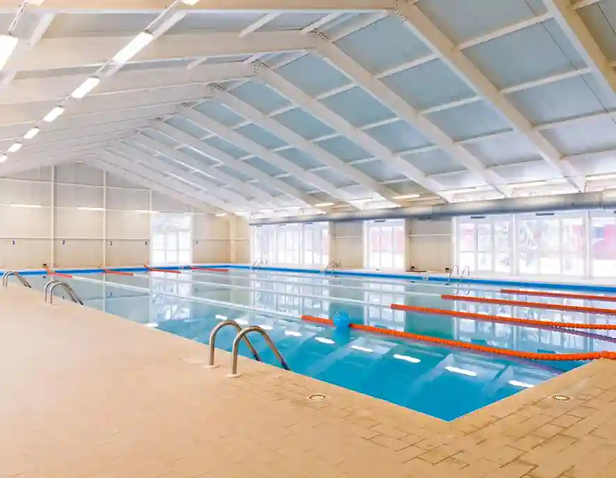 A large indoor swimming pool with clear blue water, divided by orange lane markers, under a high white ceiling with ample natural light from windows along one side. Steps lead into the pool from the tiled deck, ideal for a Residential Pool Cleaning Service to maintain.