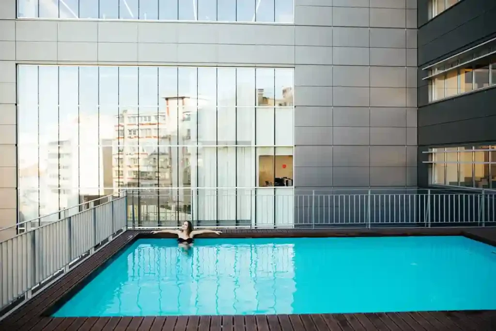 A person relaxes in a small rooftop swimming pool surrounded by a metal fence, with modern city buildings and large reflective windows in the background—an ideal spot maintained by a Commercial Pool Cleaning Service.