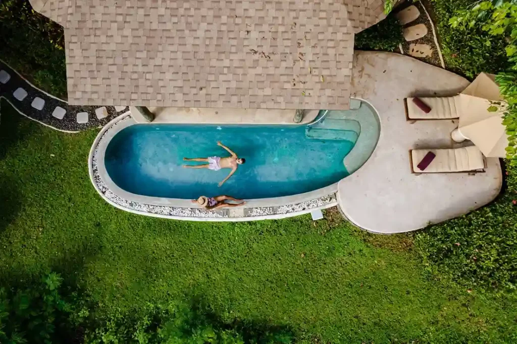 Aerial view of two people relaxing in a small, kidney-shaped pool next to a house with a shingled roof—perfect inspiration for those considering a Commercial Swimming Pool Repair Service to enhance their own backyard oasis.
