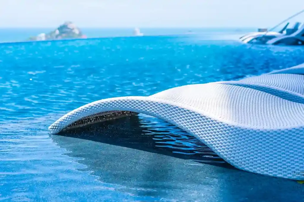 A close-up of a white woven lounge chair, part of a Commercial Spa Installation, partially submerged in the shallow edge of a blue infinity pool, with the ocean and a small island visible in the blurred background.