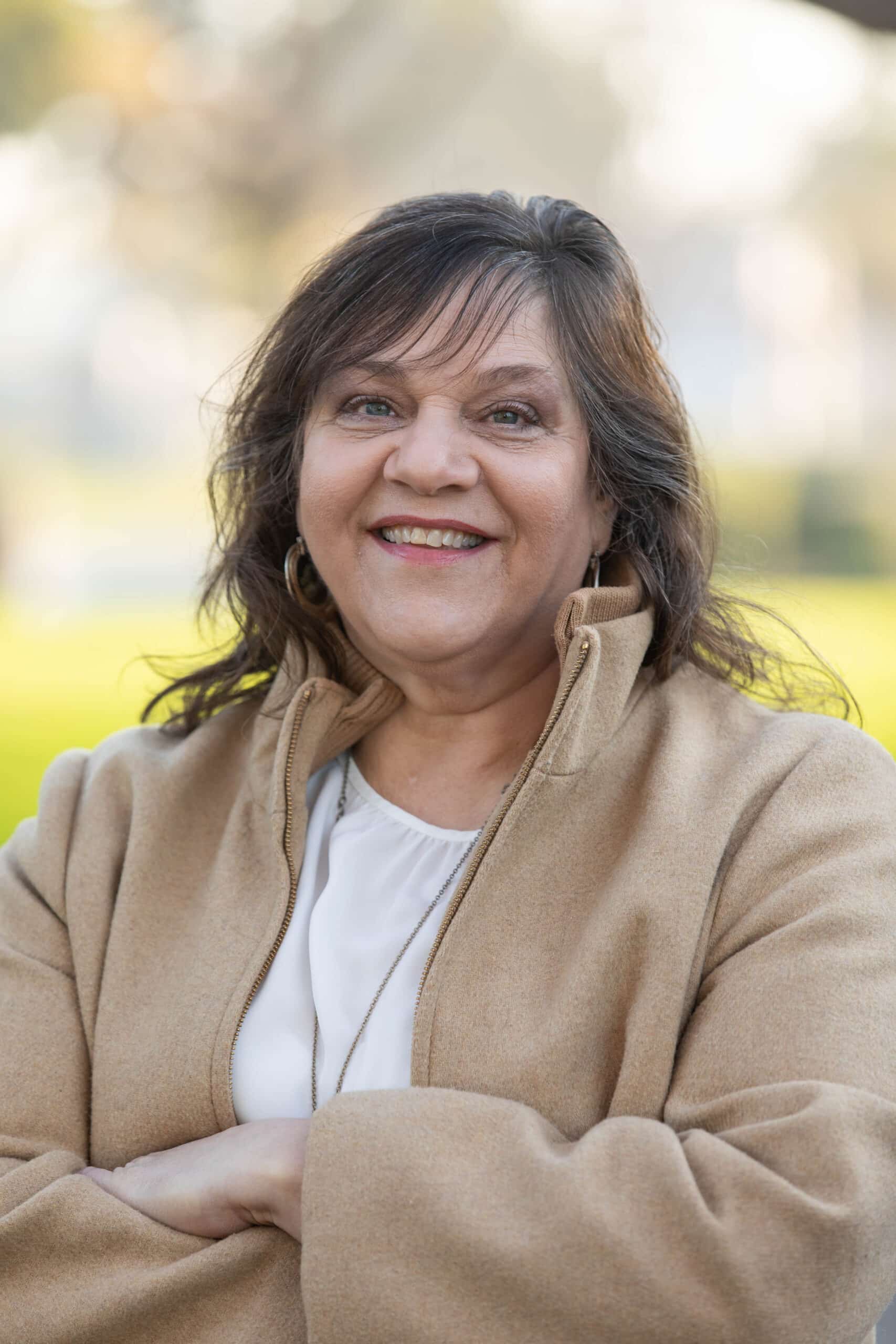 A woman with long brown hair smiles while standing outdoors in Long Beach, CA. She is wearing a tan coat over a white top and has her arms crossed. The softly blurred background hints at green and yellow tones near a swimming pool.