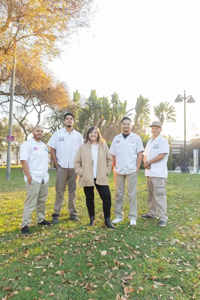 About: Five people stand outside on grass in a park; four wear matching light-colored work uniforms, while one person in the center wears a beige coat and black pants. Trees and sunlight fill the background.