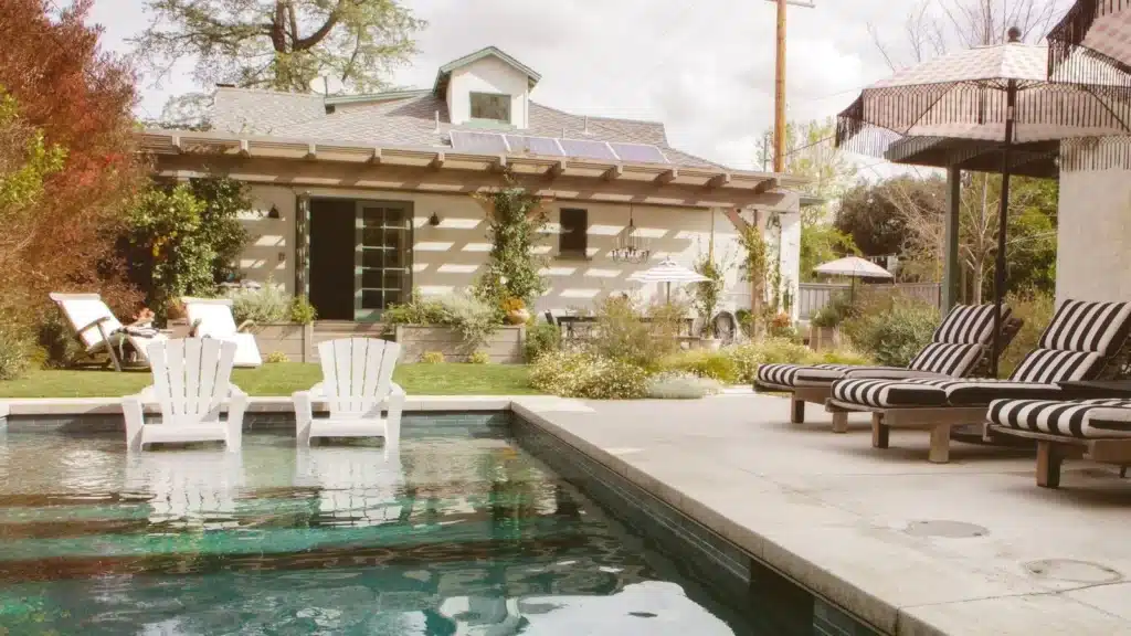 A backyard swimming pool with two white chairs in the water, striped lounge chairs and umbrellas on the poolside, and a small house with solar panels and greenery in the background, perfect for relaxing in Long Beach CA.