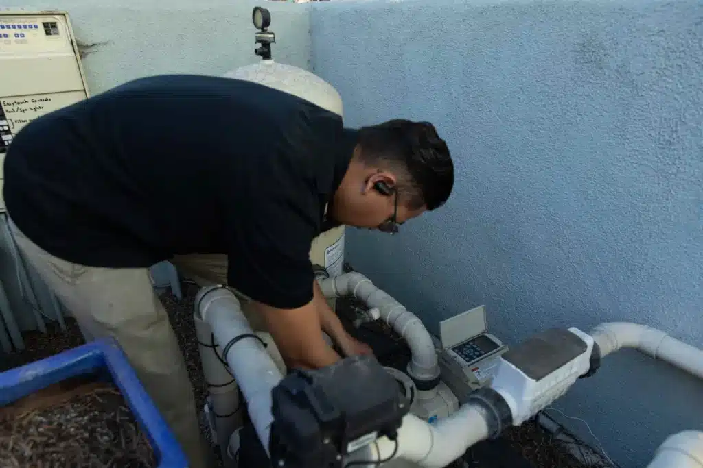 A person wearing a black shirt and earpiece bends over and works on commercial pool motor repair and pipes next to a light blue wall.