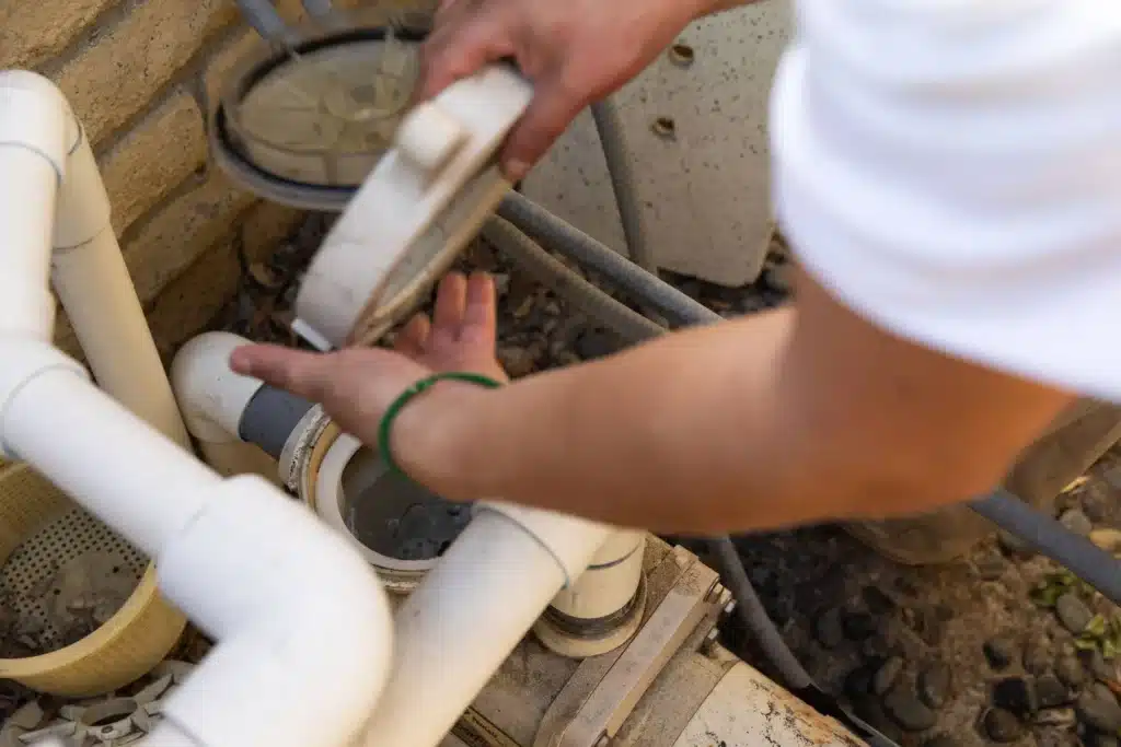 A person’s hands are cleaning or inspecting a commercial pool motor repair area, focusing on a pool filter system with white PVC pipes and a transparent filter lid, outdoors near a stone wall.