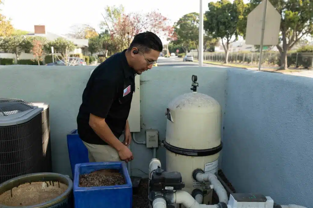 A man wearing glasses and a headset inspects commercial pool motor repair equipment and filtration systems outside near a light blue wall, with trees and a street in the background.