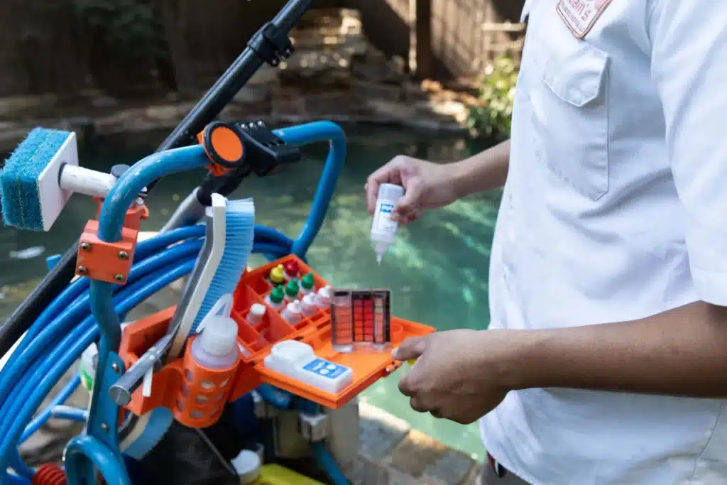 A person tests pool water using a chemical kit, holding a test tube and dropping solution, demonstrating commercial pool chemical balancing, with various pool cleaning tools and a swimming pool visible in the background.