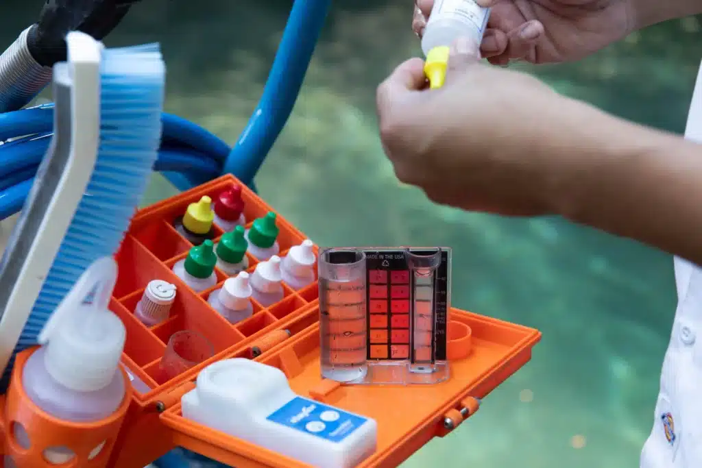 A person tests pool water using a color chart and various chemical bottles in an orange kit, demonstrating commercial pool chemical balancing, with a pool and cleaning brush visible in the background.