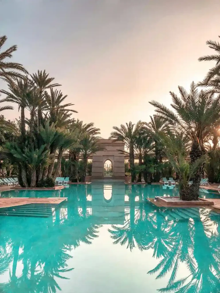A luxurious swimming pool in Long Beach, CA, is surrounded by tall palm trees, lounge chairs, and a decorative stone archway, all reflecting in the clear blue water at sunset.