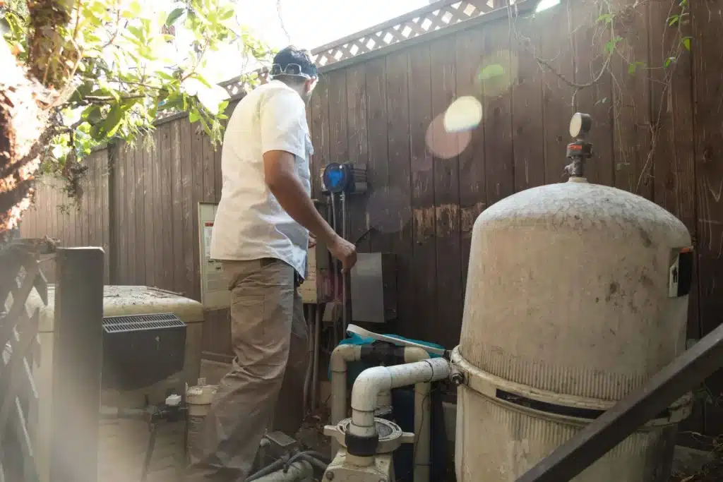 A person wearing a white shirt and beige pants is performing commercial pool filter cleaning outdoors next to a wooden fence, with sunlight shining through the trees.