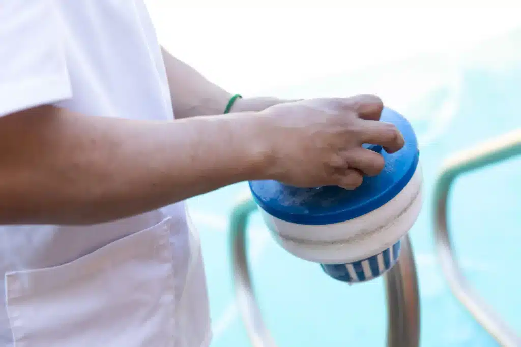 A person in a white shirt holds a blue and white pool chlorine dispenser near the edge of a swimming pool, ensuring proper sanitation before commercial pool filter cleaning.