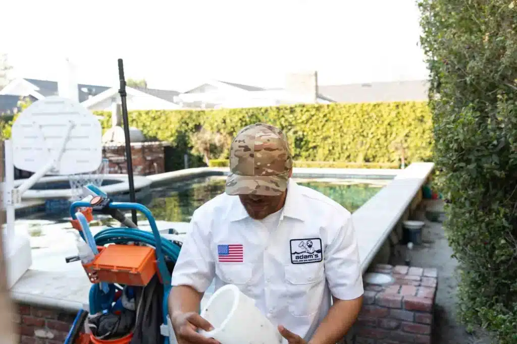 A person wearing a camouflage cap and a white shirt with an American flag patch and a pool service logo stands by a backyard pool, holding a white bucket near cleaning equipment, ready for a commercial spa installation.