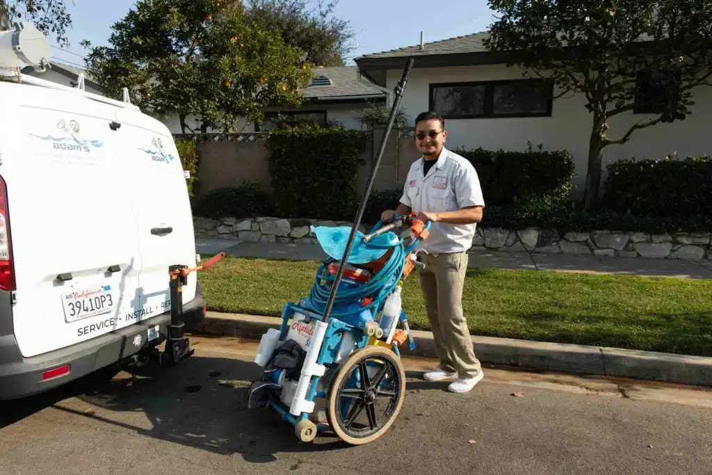 A man in a white shirt stands on a residential street beside a van, smiling and holding pool cleaning equipment on a wheeled cart—ready for commercial spa installation. Houses, trees, and bushes are visible in the background.