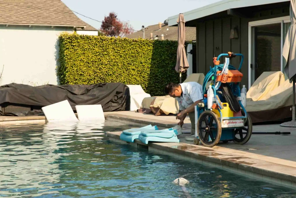 A person kneels by the edge of a backyard swimming pool, setting a blue pool wheelchair transfer device into the water—safety and accessibility features recommended by a Commercial Swimming Pool Contractor. A wheelchair with pool accessories sits nearby on the deck.