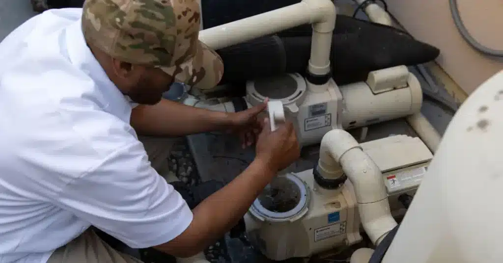 A Commercial Swimming Pool Contractor wearing a camouflage hat and white shirt is servicing a pool pump, adjusting a component on the white plumbing system next to a tan wall.