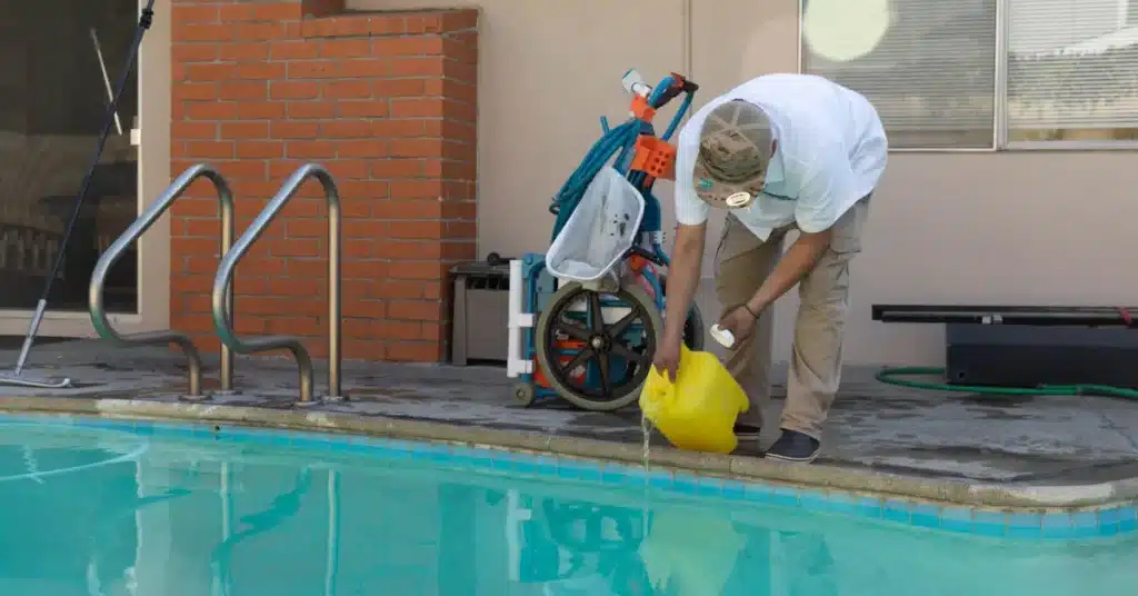 A person in a hat and white shirt, likely a commercial swimming pool contractor, pours liquid from a yellow container into a swimming pool. Pool cleaning equipment and a hose are visible nearby on the pool deck.