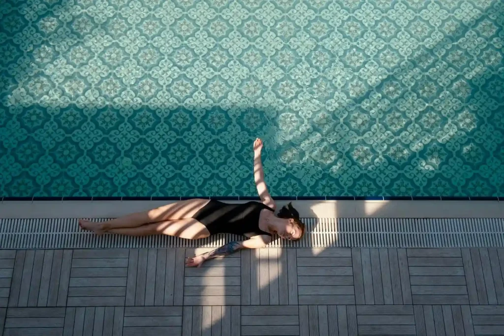 A woman in a black swimsuit lies on her back by the edge of a tiled commercial lap pool installation, with patterns of light and shadow cast across her and the pool’s surface.