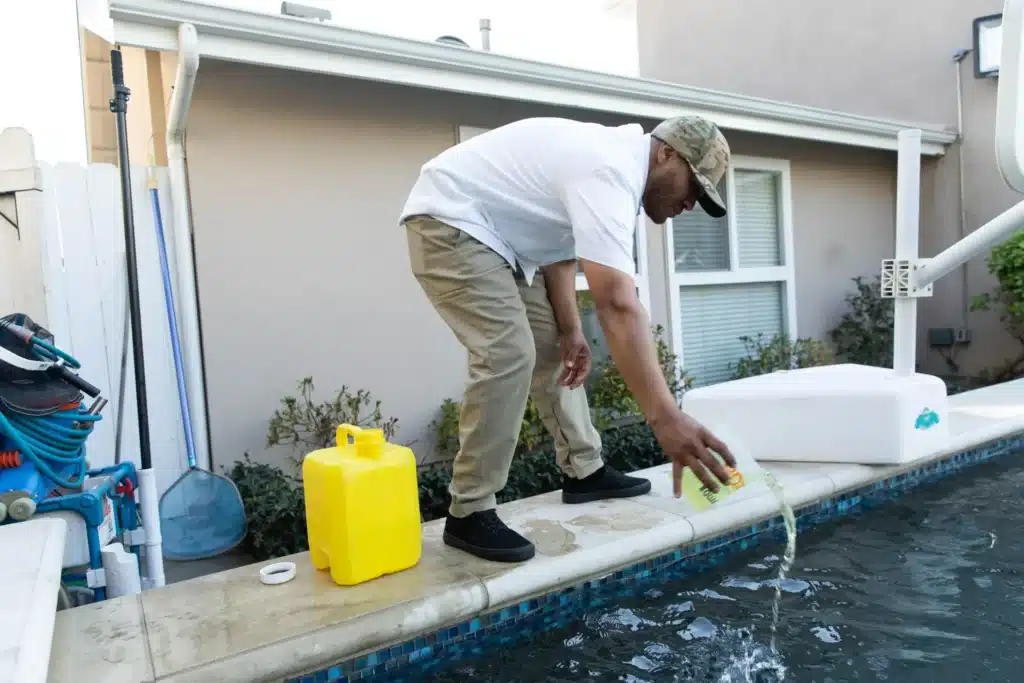 A man wearing a cap, white shirt, and khaki pants pours liquid from a yellow container into a swimming pool at a commercial pool construction site; cleaning supplies are visible nearby.