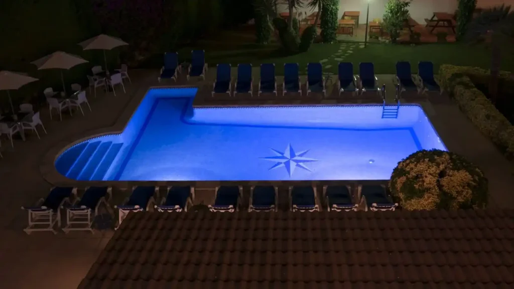 A rectangular outdoor swimming pool lit up at night, featuring a star design on the floor—perfect for commercial lap pool installation. Blue lounge chairs, umbrellas, and tables line the poolside, with lush greenery and patio furniture nearby.