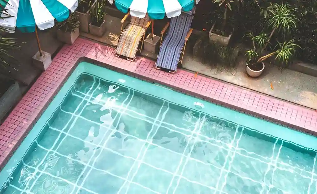 A clear outdoor swimming pool with turquoise water, crafted by a skilled swimming pool contractor, is surrounded by pink tiles. Two striped lounge chairs and a green-and-white striped umbrella sit nearby among potted plants, creating a bright and inviting scene.