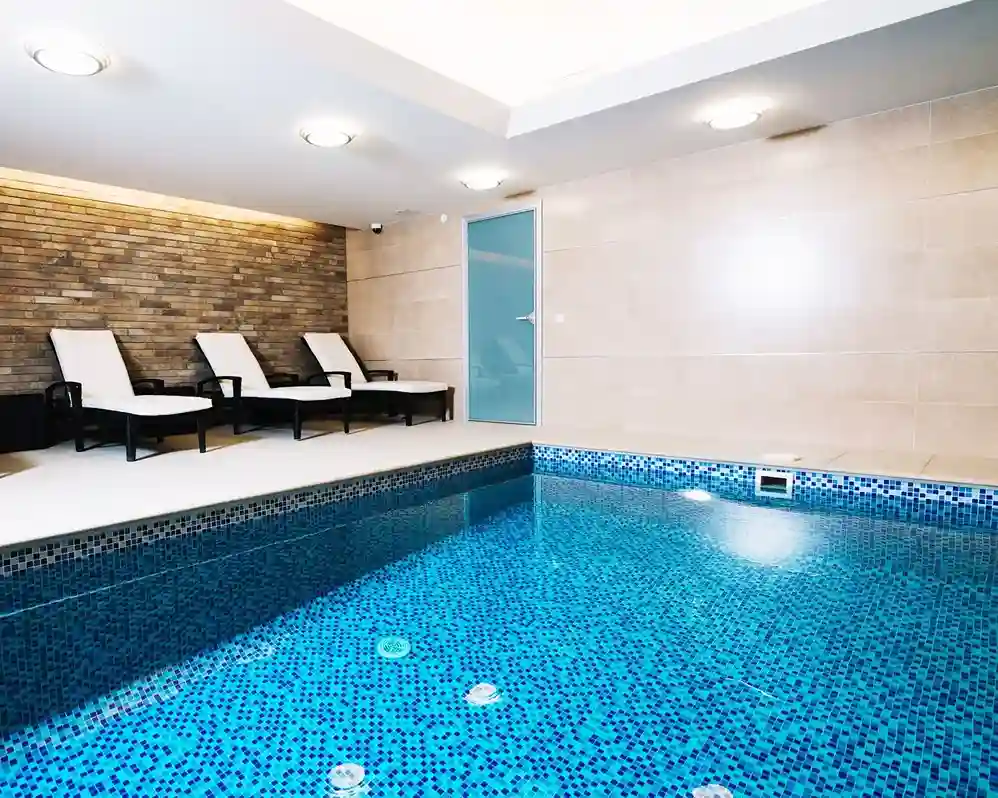 A modern indoor pool with blue mosaic tiles, this Commercial Spa Installation features beige walls, a brick accent wall, and three white lounge chairs with black frames. Ceiling lights brighten the space, while a frosted glass door adds a sleek touch.