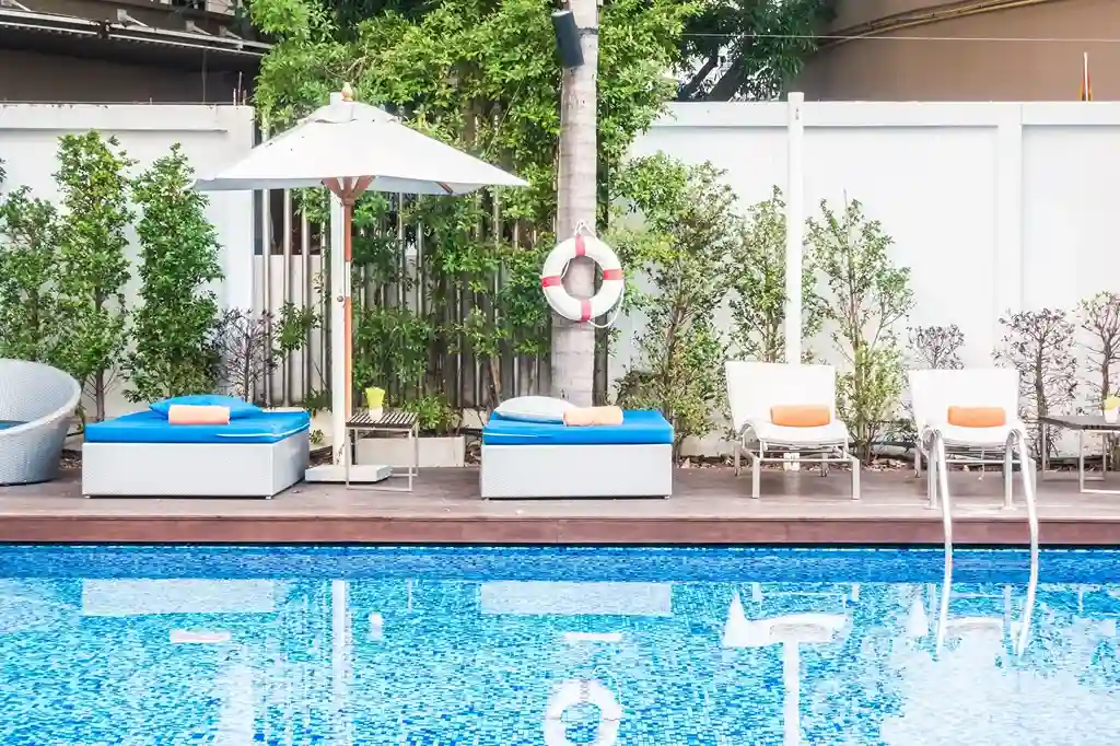 A bright outdoor pool scene by a commercial swimming pool contractor, featuring clear blue water, two lounge beds with blue cushions, a white umbrella, a lifebuoy, and white chairs, all surrounded by green plants and a white fence.