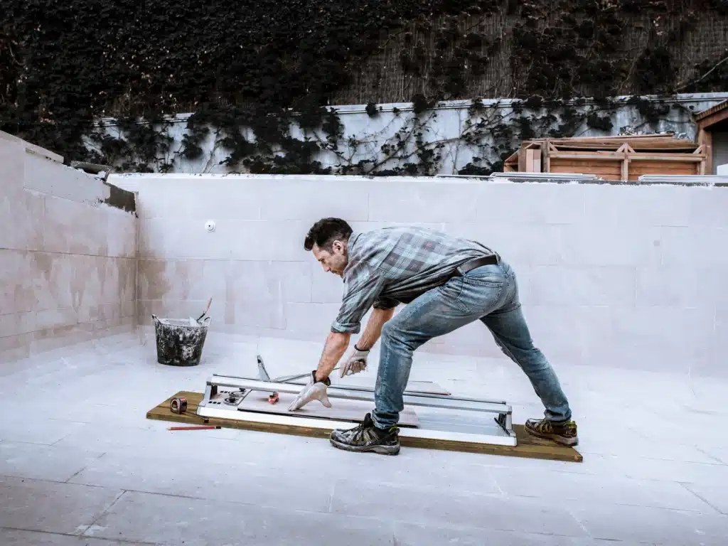 A man in jeans and a plaid shirt uses a cutting tool to work on a tile or plank in an outdoor construction area, surrounded by tools, a bucket, and building materials.