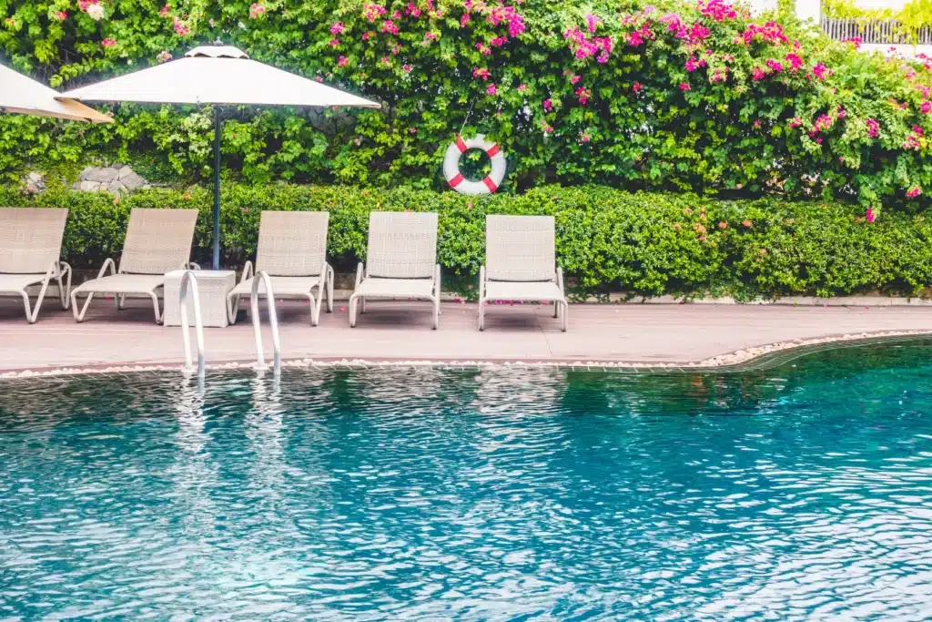 Five empty lounge chairs and two umbrellas line the edge of a swimming pool. Behind them is a lush green hedge with pink flowers and a life preserver hanging on the wall.