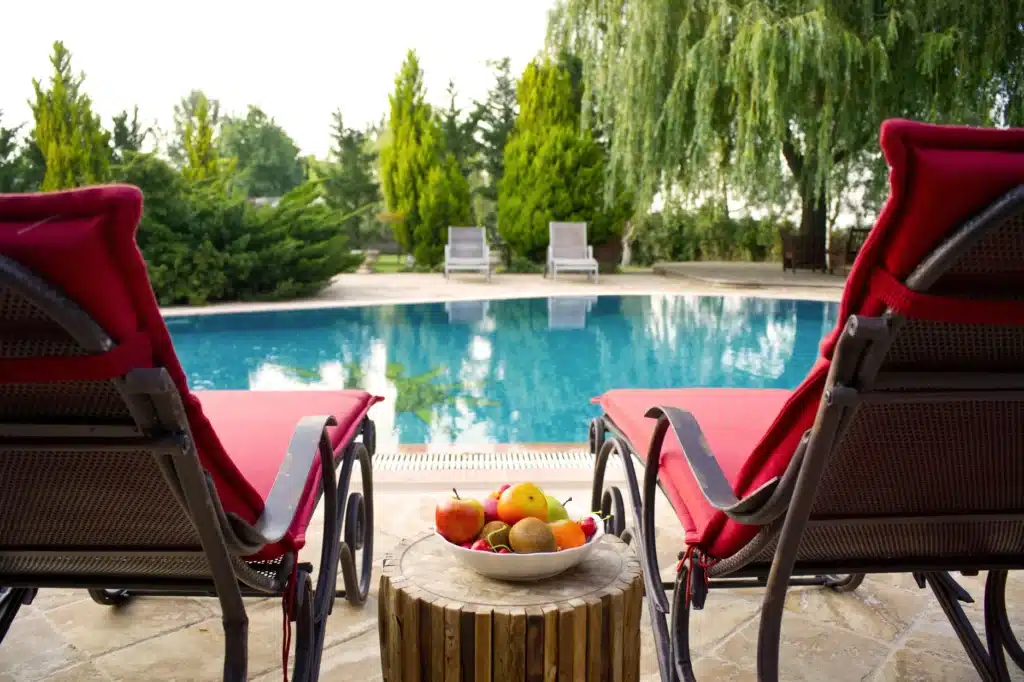 Two red lounge chairs facing a clear swimming pool, with a bowl of fruit on a small wooden table between them. Lush green trees and two empty lounge chairs are in the background.
