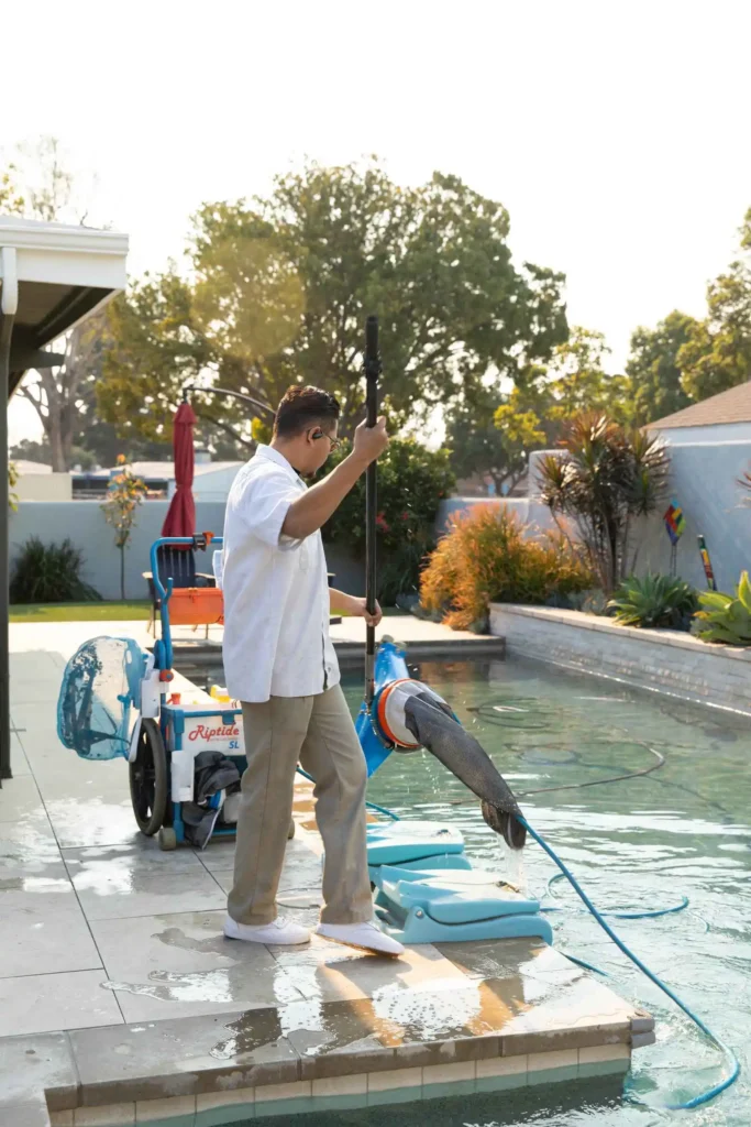 A man in a white shirt and khaki pants uses cleaning equipment to vacuum a backyard swimming pool, reflecting the care typical after a commercial lap pool installation; a pool lift chair and trees are visible in the background.