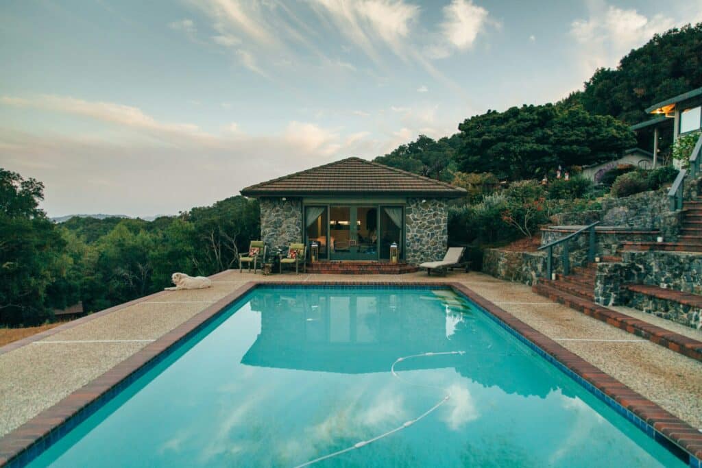 A rectangular swimming pool with clear blue water sits in front of a stone pool house in Long Beach, CA, surrounded by lush greenery and trees under a partly cloudy sunset sky. Patio furniture and a lounge chair are on the pool deck.