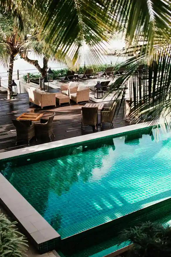 A serene swimming pool in Long Beach CA is surrounded by palm trees, with wicker chairs and tables on a wooden deck in the background, creating a relaxing tropical atmosphere.